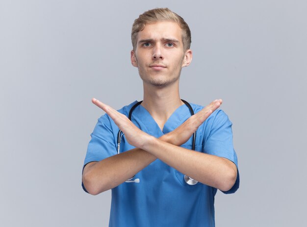Strict young male doctor wearing doctor uniform with stethoscope showing gesture of no isolated on white wall