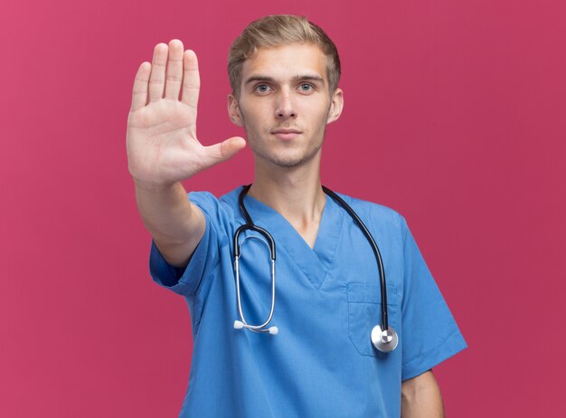 Strict young male doctor wearing doctor uniform with stethoscope showing stop gesture isolated on pink wall