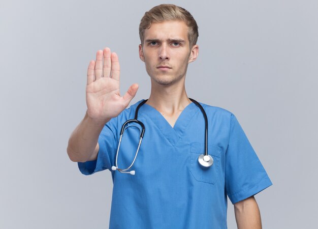 Strict young male doctor wearing doctor uniform with stethoscope showing stop gesture isolated on white wall