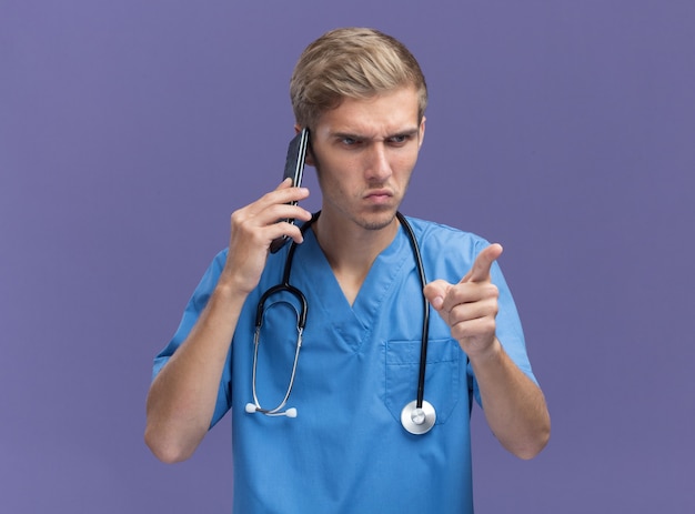 Strict young male doctor wearing doctor uniform with stethoscope speaks on phone points at side isolated on blue wall