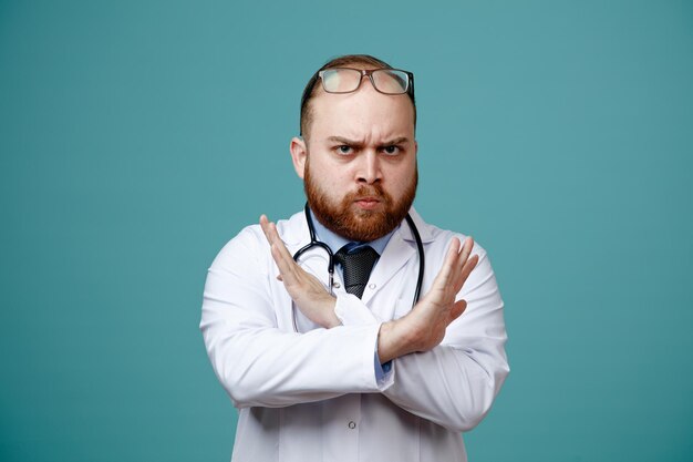 Strict young male doctor wearing medical coat and stethoscope around his neck and glasses on head looking at camera showing no gesture isolated on blue background