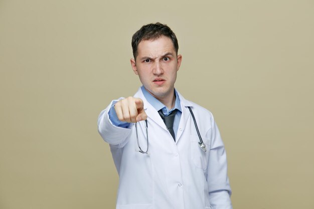 Строгий молодой врач-мужчина в медицинском халате и стетоскопе на шее смотрит и указывает на камеру, изолированную на оливково-зеленом фоне