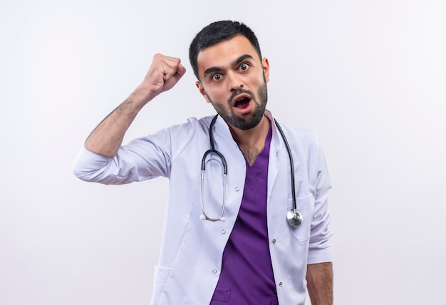 Strict young male doctor wearing stethoscope medical gown raising fist on isolated white background