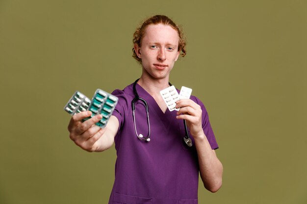 Strict young male doctor wearing uniform with stethoscope holding pills isolated on green background