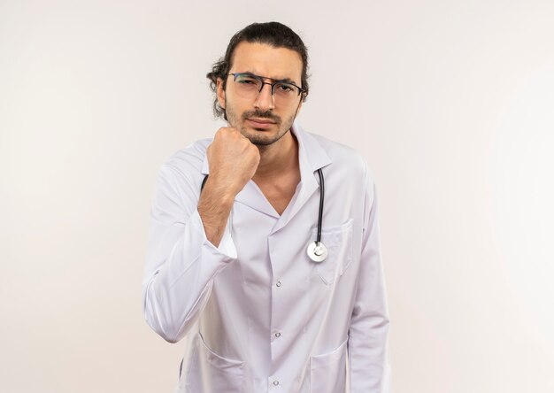 Strict young male doctor with optical glasses wearing white robe with stethoscope raising fist on isolated white wall with copy space