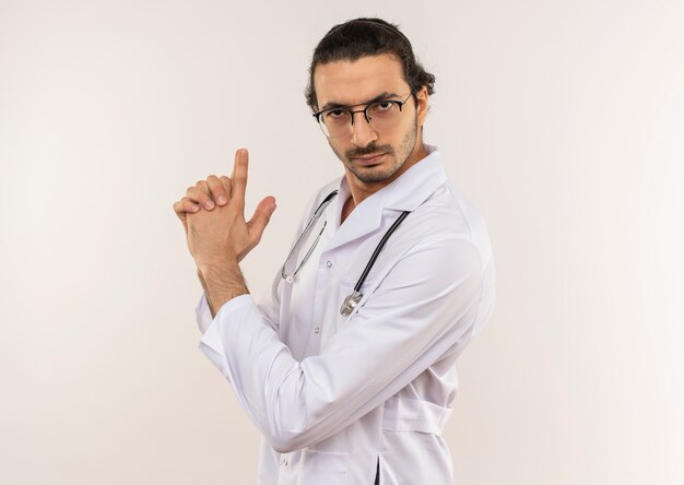 Strict young male doctor with optical glasses wearing white robe with stethoscope sgowing pistol gesture on isolated white wall with copy space