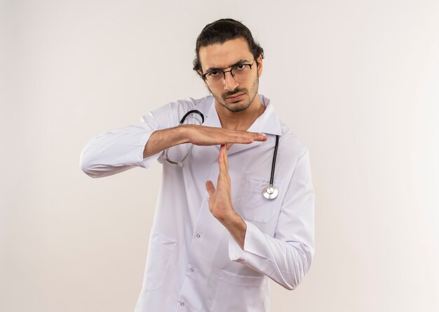 Strict young male doctor with optical glasses wearing white robe with stethoscope showing timeout gesture on isolated white wall with copy space