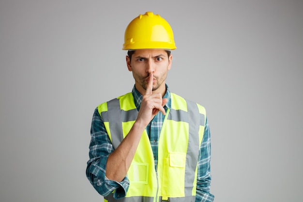 Бесплатное фото Строгий молодой инженер-мужчина в защитном шлеме и униформе смотрит в камеру, показывая жест молчания на белом фоне