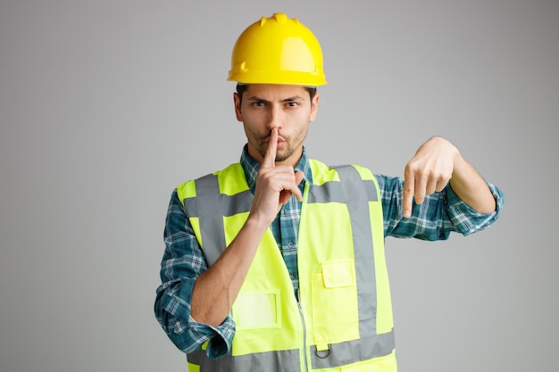 Strict young male engineer wearing safety helmet and uniform looking at camera showing be silent gesture pointing down isolated on white background