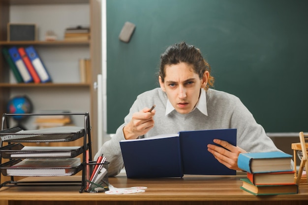 strict young male teacher holding notebbok with pencil sitting at desk with school tools on in classroom