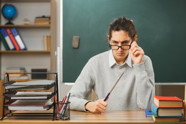 строгий молодой учитель-мужчина в очках, держащий указатель, сидящий за партой со школьными инструментами в классе