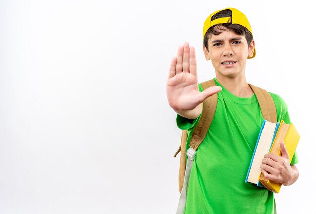 Strict young school boy wearing backpack with cap holding books showing stop gesture 