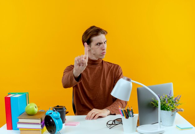 Strict young student boy sitting at desk with school tools showing one isolated on yellow wall