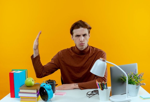 Strict young student boy sitting at desk with school tools showing stop gesture out to side on yellow