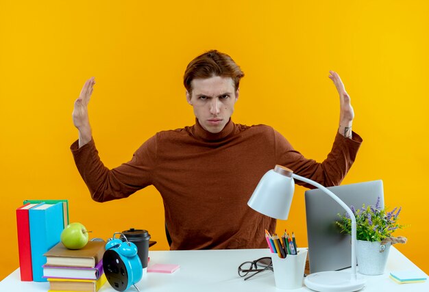 Strict young student boy sitting at desk with school tools showing stop gesture to side on yellow