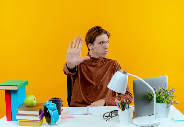 Strict young student boy sitting at desk with school tools showing stop gesture on yellow