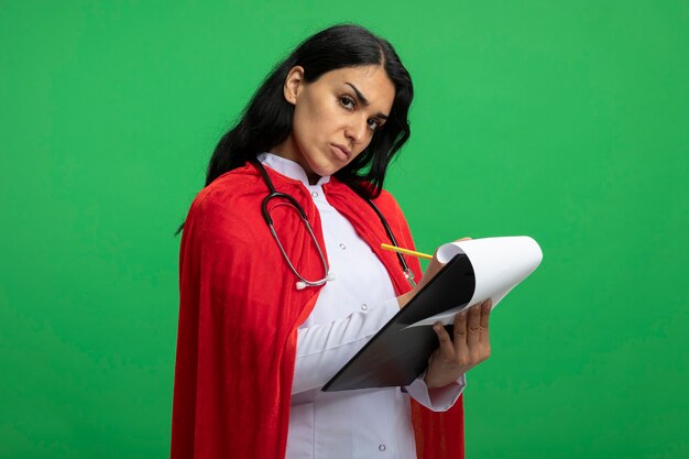 緑に分離されたクリップボードに聴診器を持って何かを書いている医療ローブを着てまっすぐ前を見てまっすぐに見ている厳格な若いスーパーヒーローの女の子