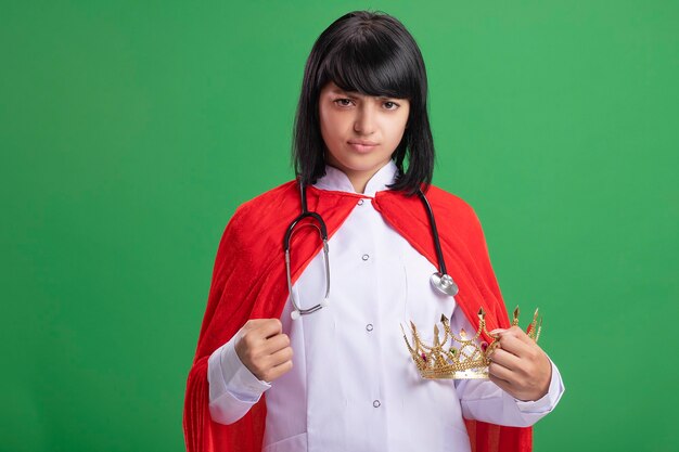 Free photo strict young superhero girl wearing stethoscope with medical robe and cloak holding crown isolated on green wall