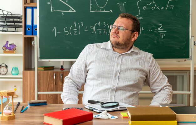 strict young teacher wearing glasses sitting at desk with school supplies in classroom keeping hand on waist looking at side