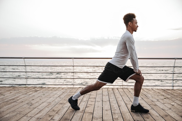 Strong active curly dark-skinned man in black shorts and white long-sleeved t-shirt squats, works out and stretches near sea