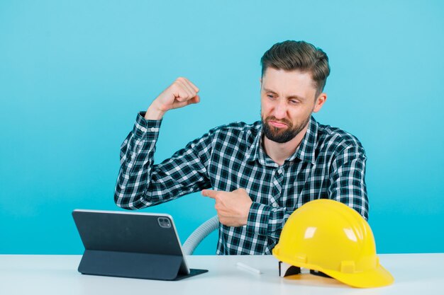 Strong architect is showing his muscle by sitting in front of tablet on blue background