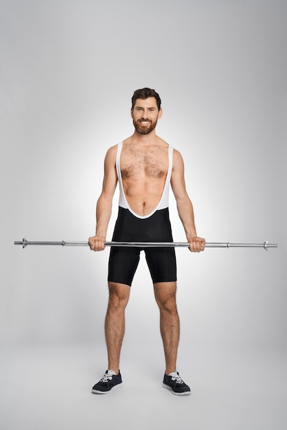 Free photo strong bearded powerlifter posing with unweighted barbell in studio front view of smiling male