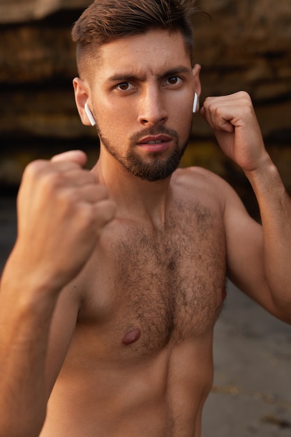 Сильный боксер с обнаженным телом, сжимает кулаки, выполняет упражнения на удары ногами, тренирует бокс на открытом воздухе.
