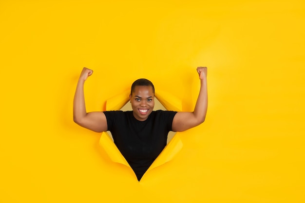 Free photo strong. cheerful african-american young woman poses in torn yellow paper, emotional and expressive.