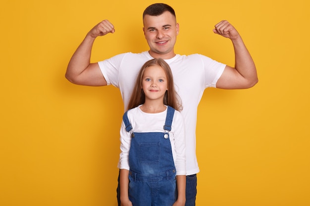 сильный отец, показывающий бицепс, притворяющийся сильным, одетый в белую повседневную футболку, мотивированную на успешное будущее, стоит на желтой стене с его дочерью, одетой в джинсовые туфли.