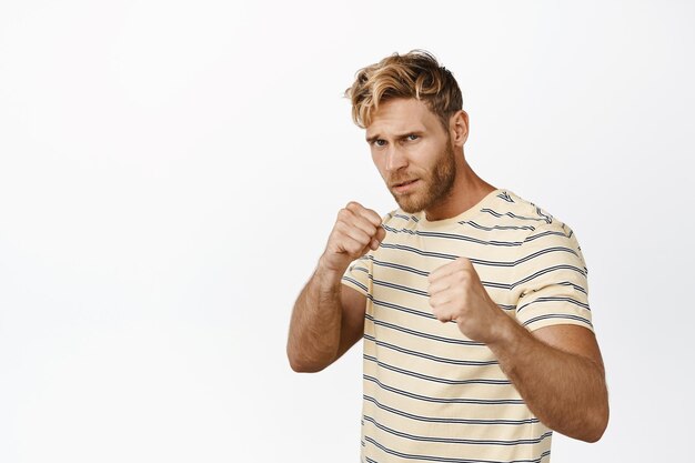 Free photo strong handsome blond man standing in boxing fighter pose clenching fists ready for fight posing in casual tshirt over white background