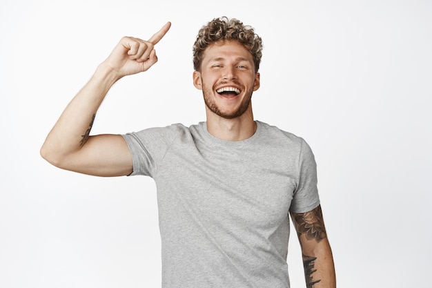 Strong handsome guy laughing flexing biceps showing muscle on arm and pointing finger up showing sport related advertisement standing over white background