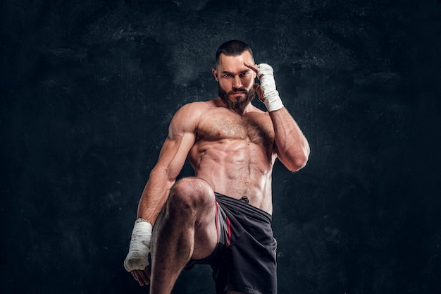 Strong muscular fighter is showing his punch while posing for photographer at dark photo studio.