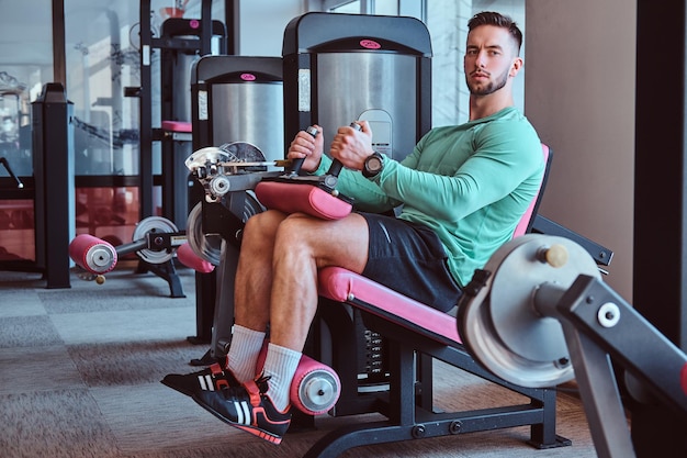 강한 생각에 잠겨있는 남자가 체육관에서 훈련 기구에 앉아 다리 운동을 하고 있습니다.