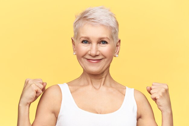 Strong positive middle aged woman with dyed short hair clenching fists, showing biceps, posing isolated. Blonde mature female having confident proud look.