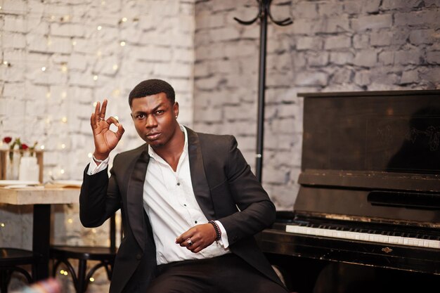 Strong powerful african american man in black suit sitting against piano and show ok sign