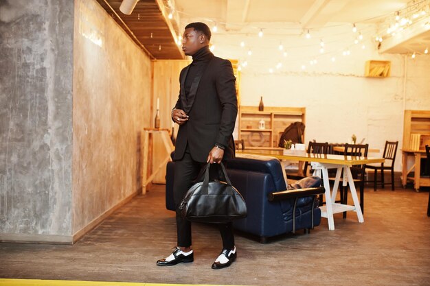 Strong powerful african american man in black suit and turtleneck with handbag posing in office