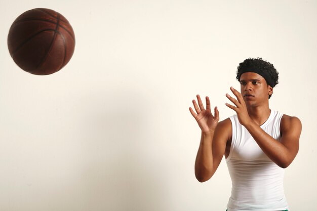 Сильный серьезный темнокожий спортсмен с афро в простой белой рубашке без рукавов, бросающий темно-коричневый винтажный баскетбольный мяч на белый
