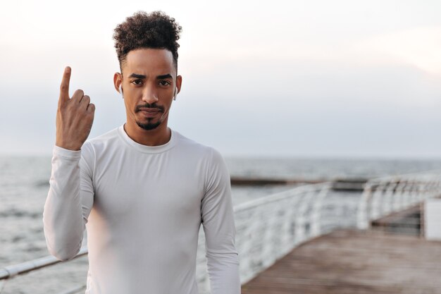 Strong serious curly brunette man in long-sleeved white t-shirt listens to music in headphones and points up near sea