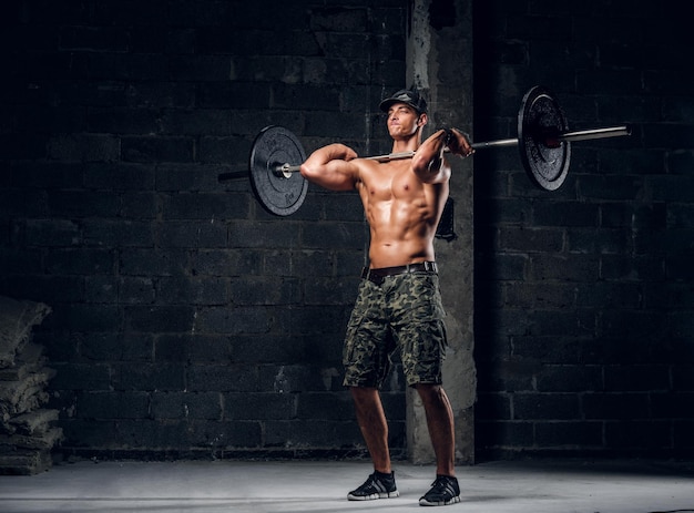 Strong shirtless man in cap is lifting barbell at dark photo studio.