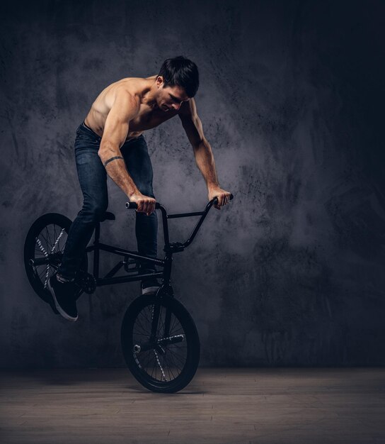 Strong shirtless man, makes an acrobatic figure with BMX in a studio. Isolated on a dark background.