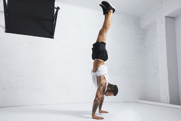 Strong tattooed in white unlabeled tank t-shirt male athlete shows calisthenic moves Handstand position