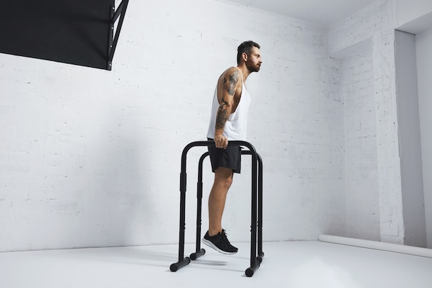 Forte tatuato in t-shirt da carro armato bianco senza etichetta, atleta maschio mostra movimenti calistenici tenendo la posizione sulle barre parallele prima dei tuffi classici, guardando dritto