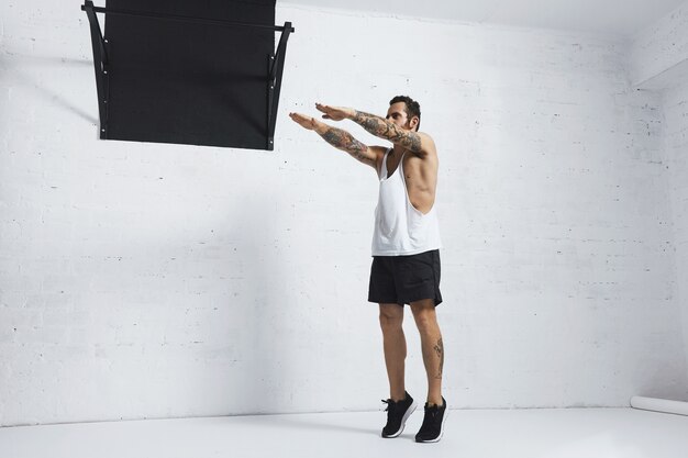 Strong tattooed in white unlabeled tank t-shirt male athlete shows calisthenic moves Squat calf rises, top position