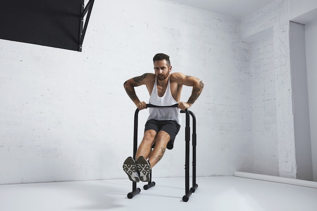 Strong tattooed in white unlabeled tank t-shirt male athlete shows calisthenic moves Straight dips move two, dip in on one of parallel bars