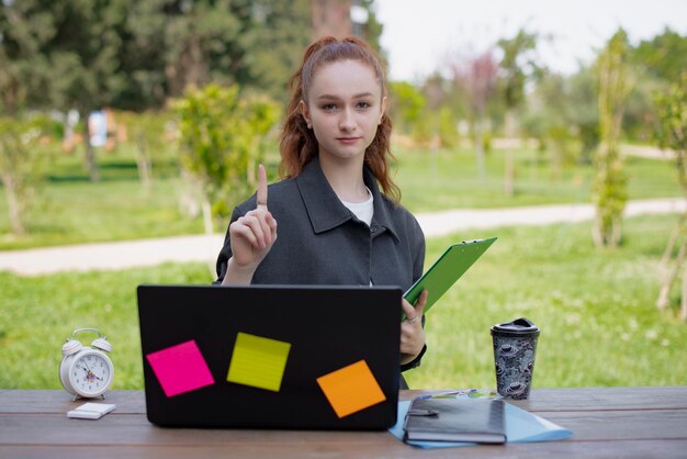 Student freelancer working at laptop in park raising finger