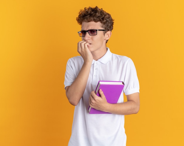 Студент парень в повседневной одежде в очках держит книгу, глядя в камеру, напряженно и нервно кусая ногти, стоя на оранжевом фоне