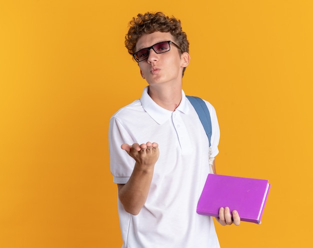 Student guy in casual clothing wearing glasses with backpack holding book blowing a kiss holding hand in front of him standing over orange background