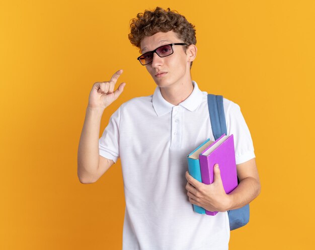 Студент парень в повседневной одежде в очках с рюкзаком держит книги, глядя в камеру, показывает жест маленького размера с пальцами, стоящими на оранжевом фоне