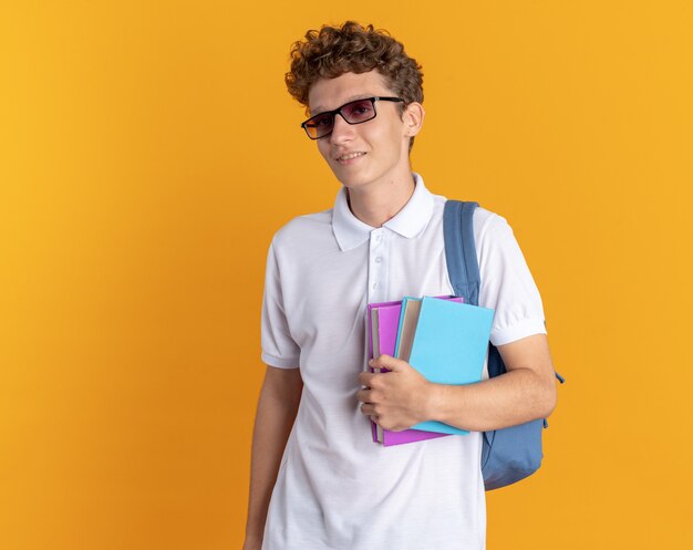 Student guy in casual clothing wearing glasses with backpack holding books looking at camera smiling confident