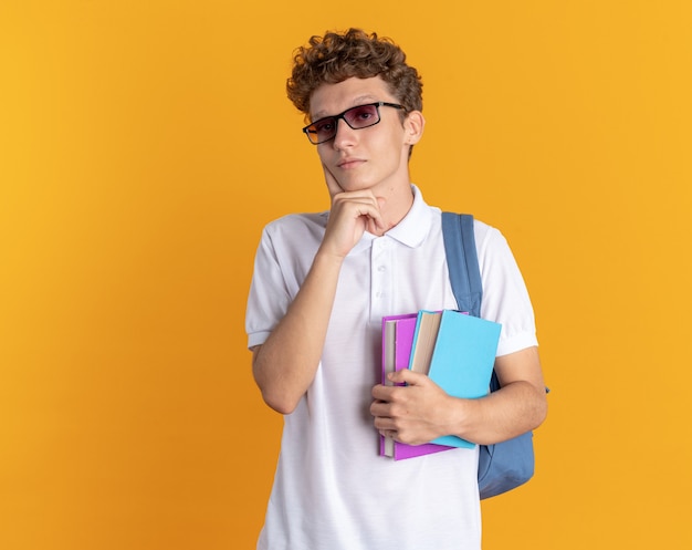 Student guy in casual clothing wearing glasses with backpack holding books looking at camera